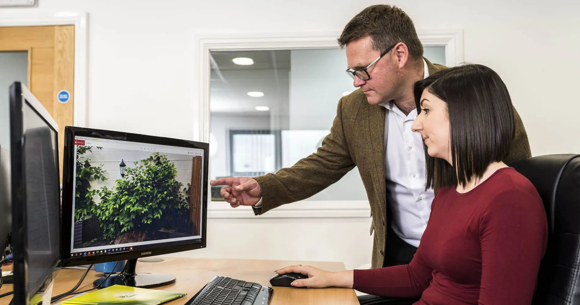 Man in a brown blazer points at a computer screen while a woman sits at a desk watching the monitor in an office setting.
