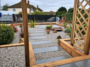 pebble stepping stones webp | Blue Iris Landscapes | Blue Iris Landscapes Residential garden with a gravel yard, stepping stone path, and wooden pergola framing a circular metal fire pit.