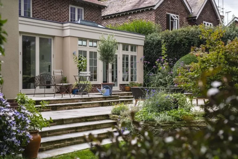 Summer-House-WEBP 2 | Blue Iris Landscapes | Blue Iris Landscapes Stone patio with steps leading to a glass-door extension, surrounded by potted plants and flowering shrubs in a sunny backyard garden.
