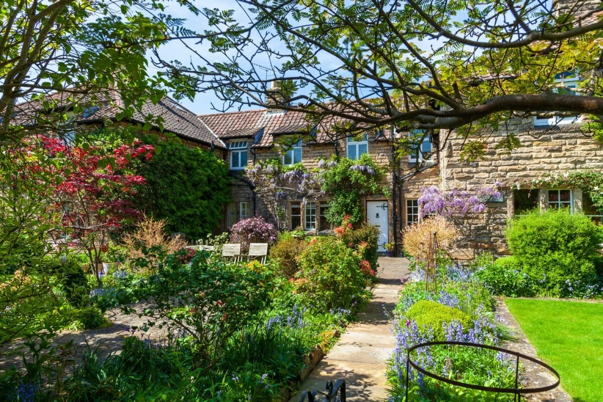 Stone cottage with a lush flowering garden and stone path leading to a white door, framed by blossoming vines.