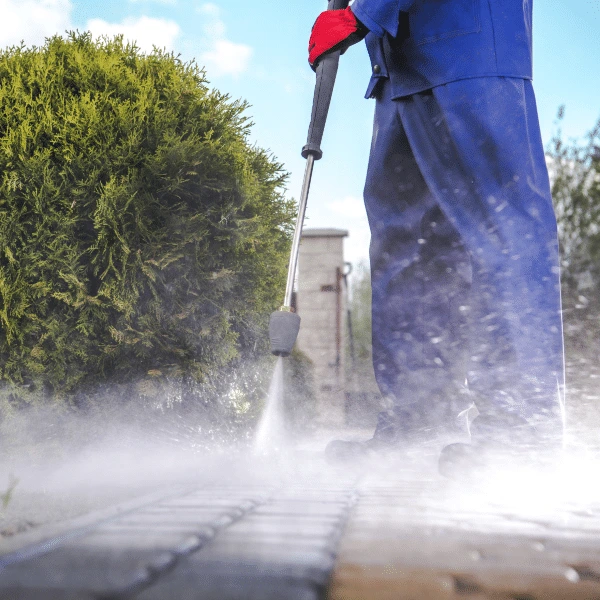 Person in blue workwear and red gloves using a pressure washer to clean a brick walkway, with spray creating mist near a trimmed hedge.