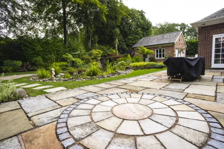 stone paving webp | Blue Iris Landscapes | Blue Iris Landscapes Circular stone patio with a concentric design, bordered by blue-gray stones, in a landscaped backyard garden with greenery and a brick house in the background. A covered grill sits on the patio to the right.