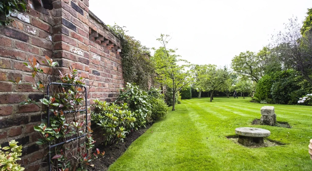 Brick garden wall on the left with climbing plants, a neatly mown lawn, and trees in the distance.